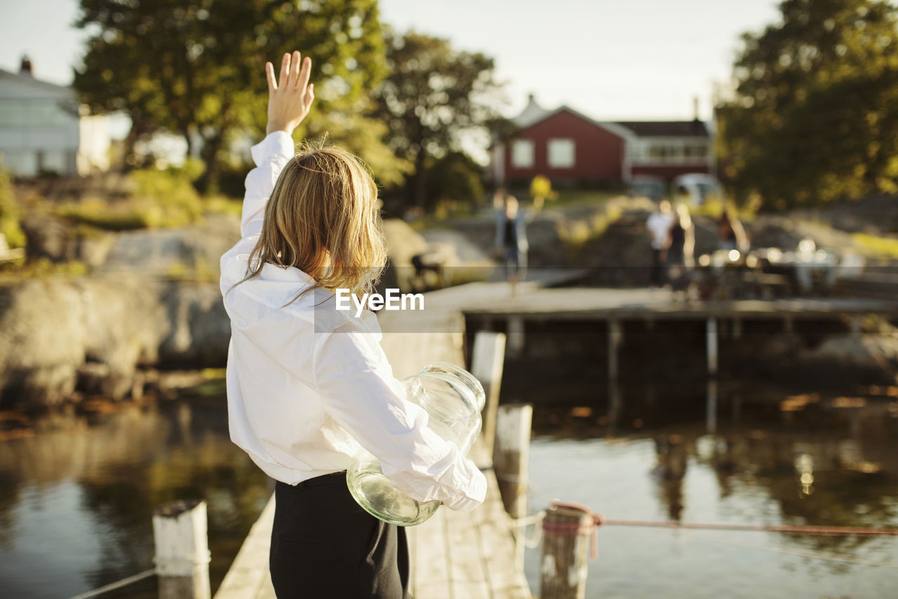 Rear view of young woman waving to friends standing on jetty at harbor