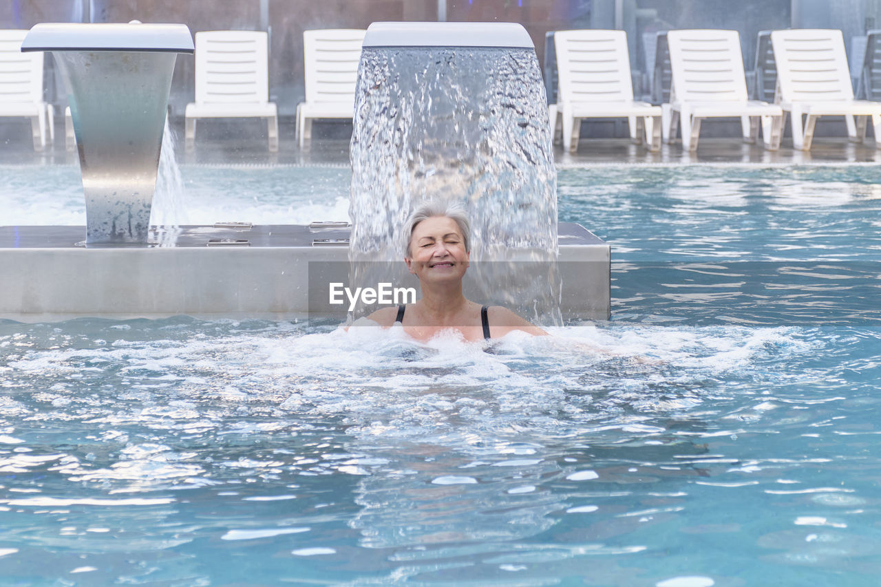 Senior caucasian smiling woman enjoying falling jets of water. hydromassage in outdoor thermal  pool