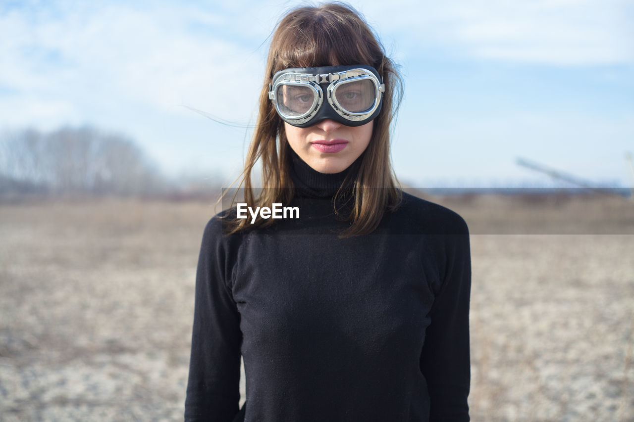 Portrait of young woman wearing swimming goggles while standing on field against sky