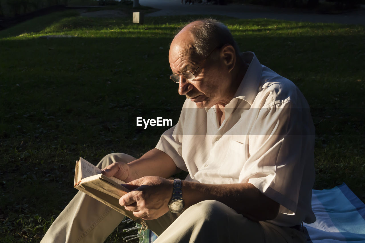 An elderly man with a watch on his hand and glasses is reading a book in a park in the fresh air.