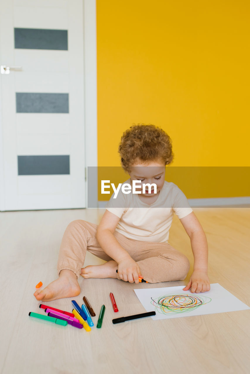 Cute little boy is sitting at home on the floor and drawing a drawing with colored pencil