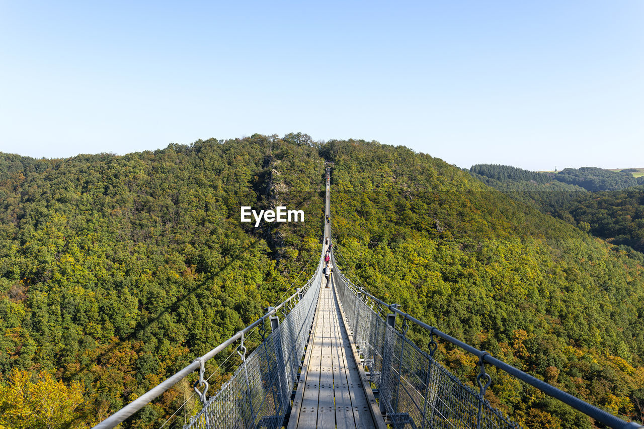 Suspension wooden bridge with steel ropes over a dense forest in west germany, visible tourists. 