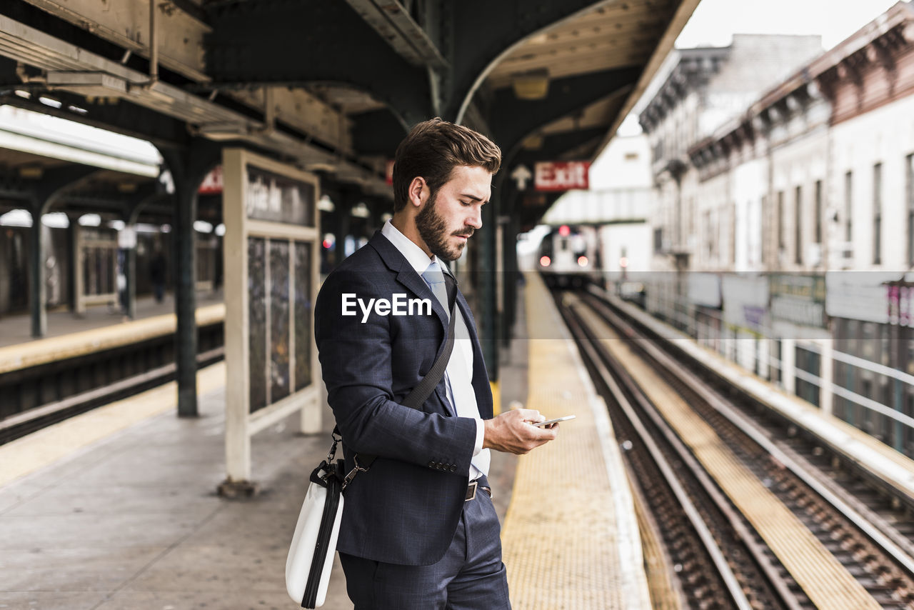 Young businessman waiting at metro station platform, using smart phone