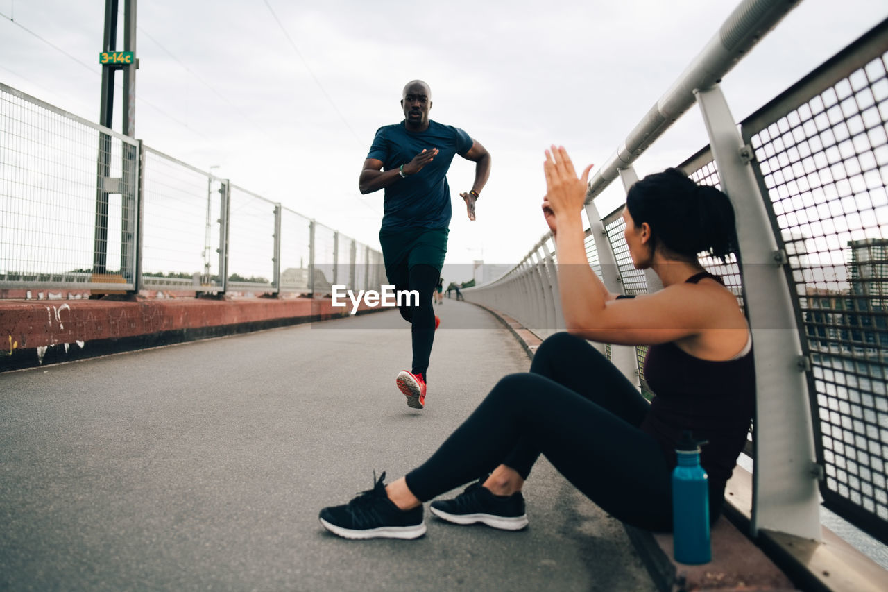 Sportswoman cheering male athlete running on footbridge in city