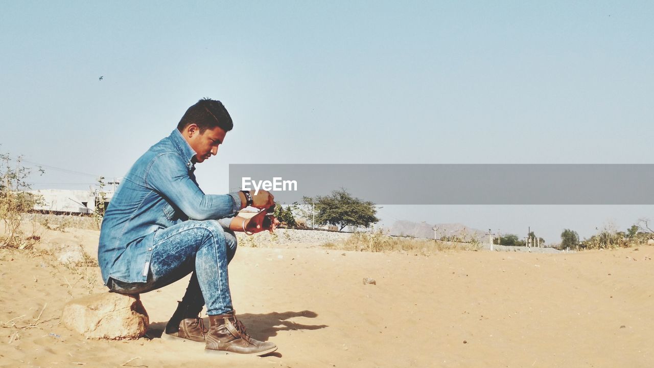 Full length side view of man holding sunglass on sandy field during sunny day