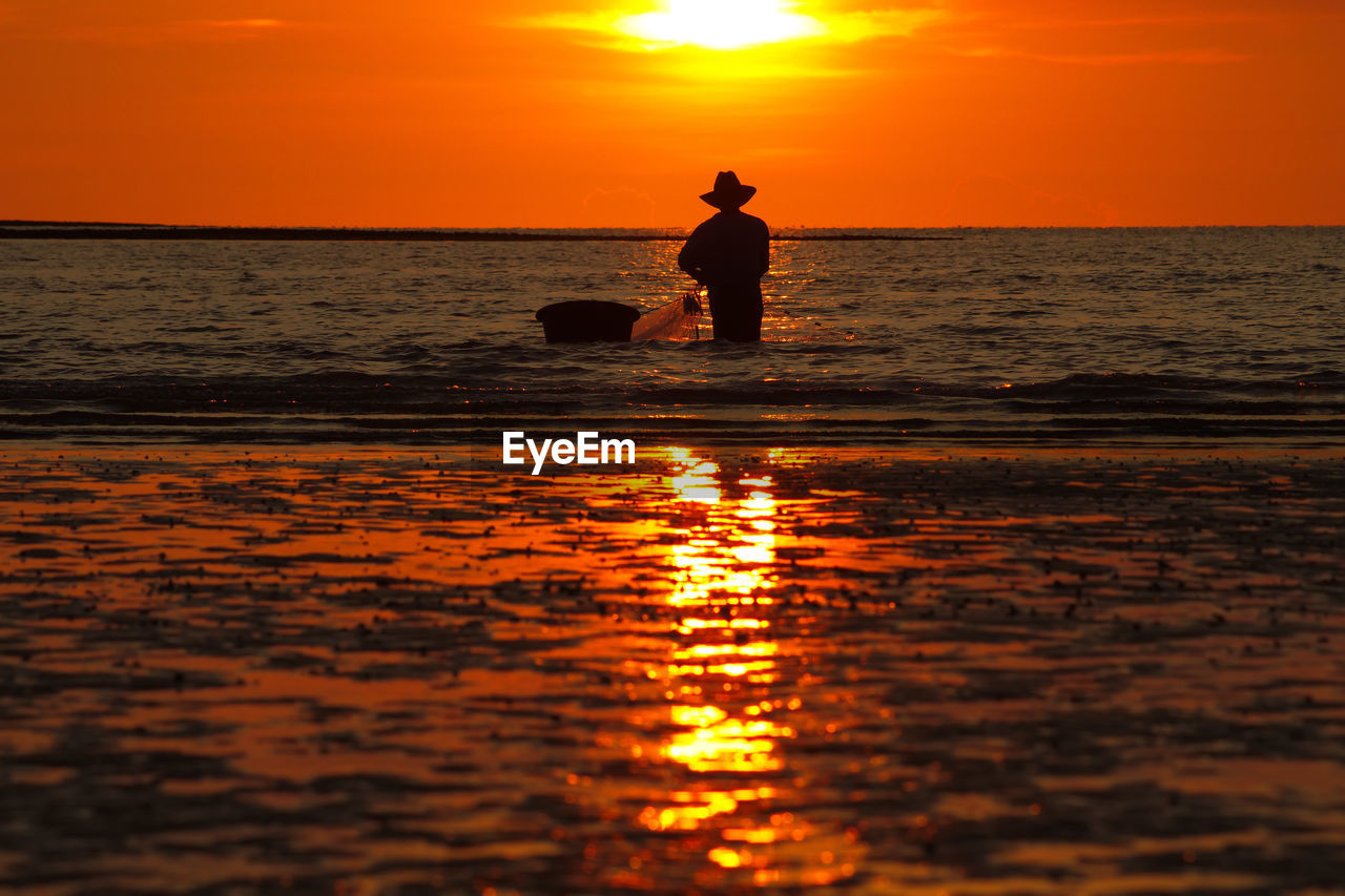 Silhouette fisherman fishing in sea against sky during sunset