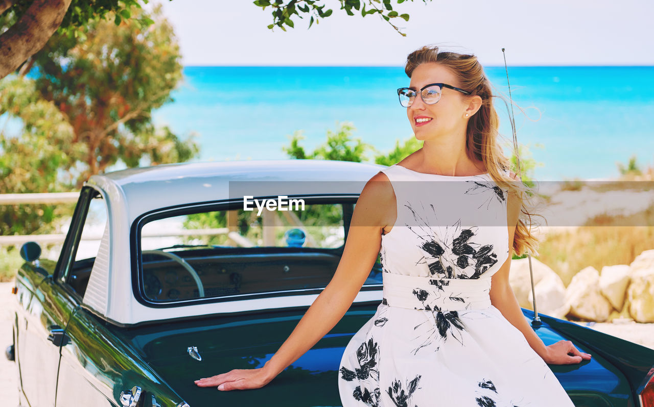 Mid adult woman sitting on vintage car against sea