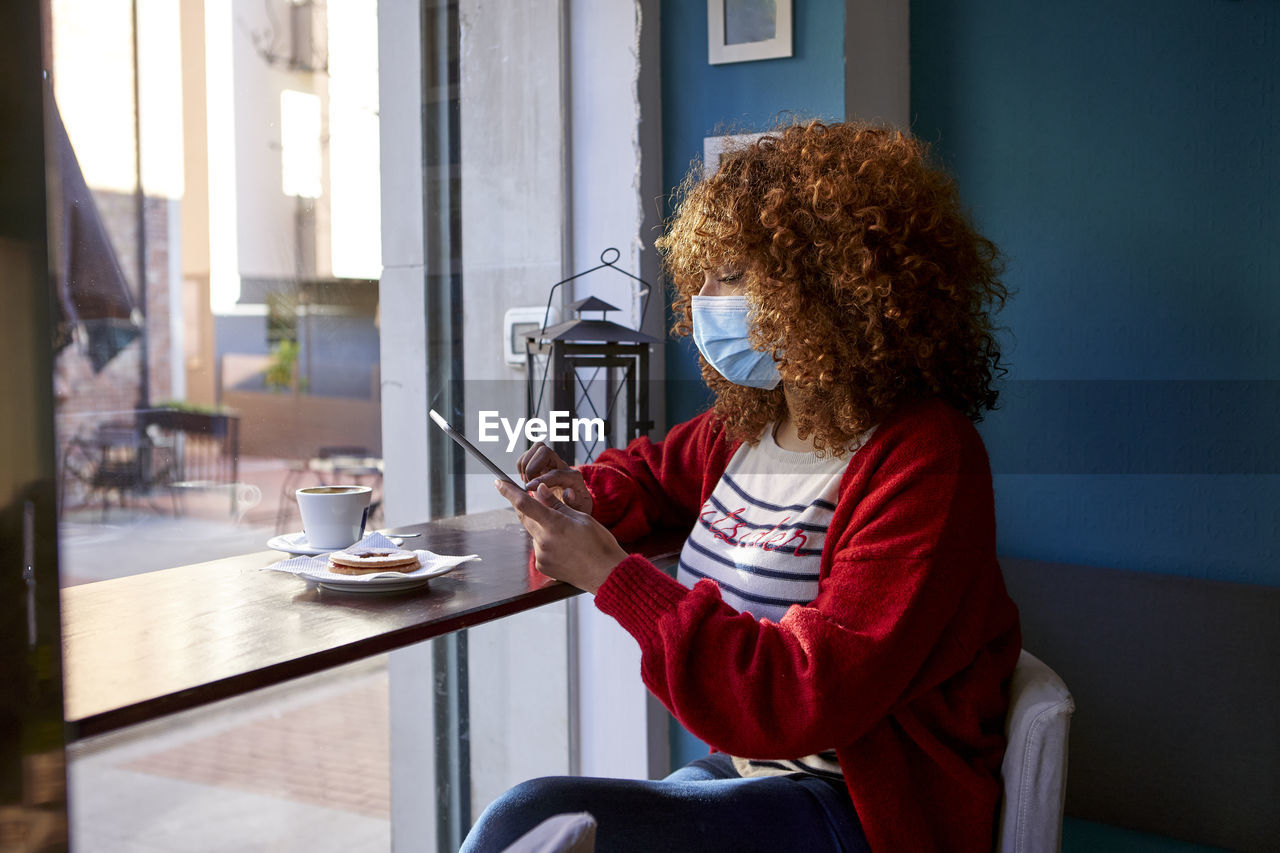 Afro woman wearing sanitary mask using digital tablet while sitting at cafe