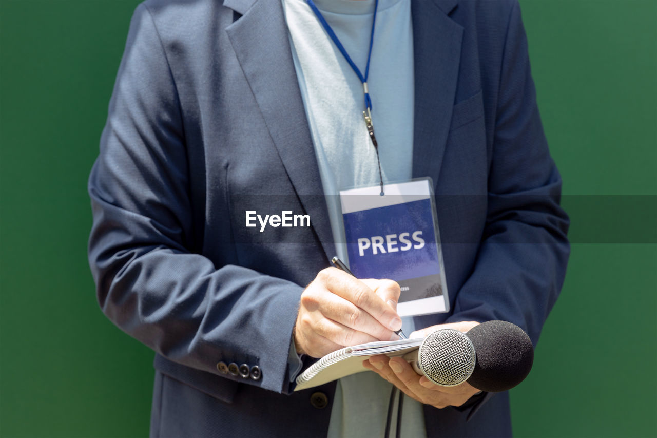 Journalist with press pass at news conference or media event, holding microphone, writing notes