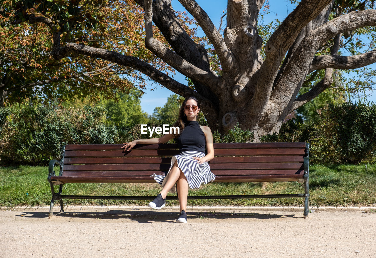 bench, tree, full length, seat, one person, plant, sitting, park bench, park, adult, park - man made space, furniture, nature, women, young adult, relaxation, leisure activity, casual clothing, lifestyles, day, footwear, sunlight, front view, portrait, outdoors, spring, emotion, fashion, vacation, outdoor furniture, person, shadow, clothing, hairstyle, female, flower, trunk, tree trunk, sunny, happiness, looking at camera, smiling, tranquility, looking