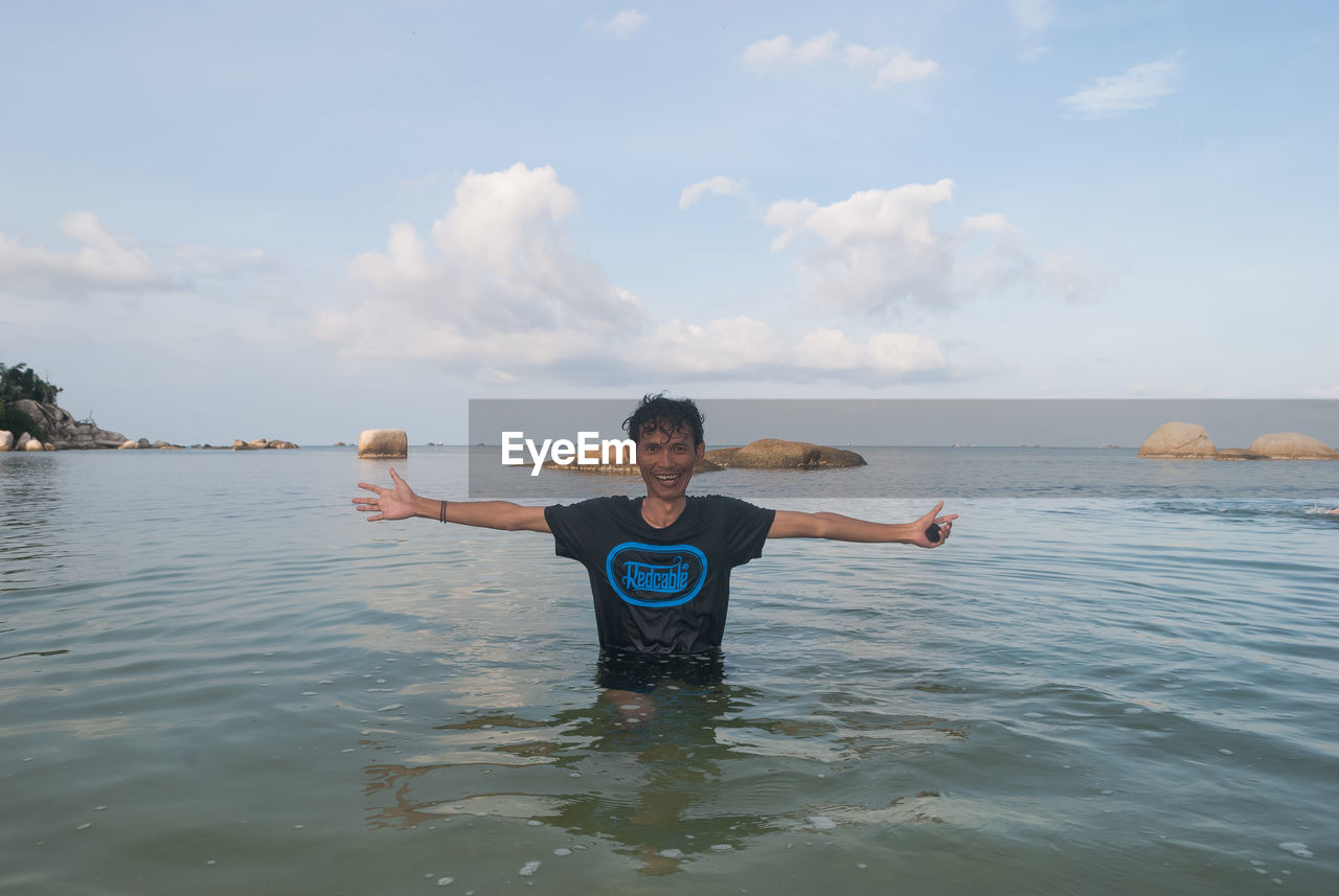 Portrait of mid adult man with arms outstretched standing in sea against sky