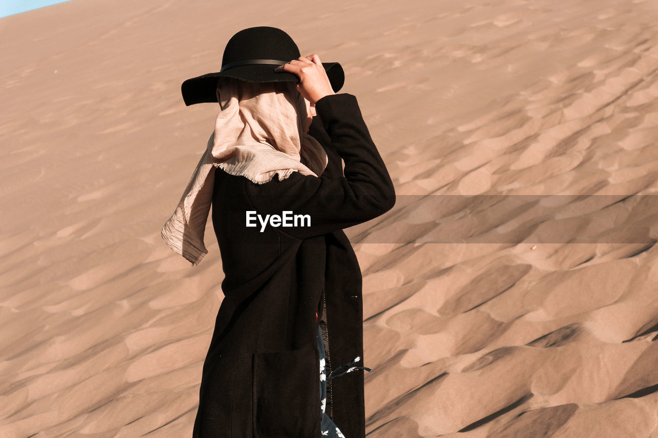 Side view of woman wearing hat and headscarf standing on sand dunes