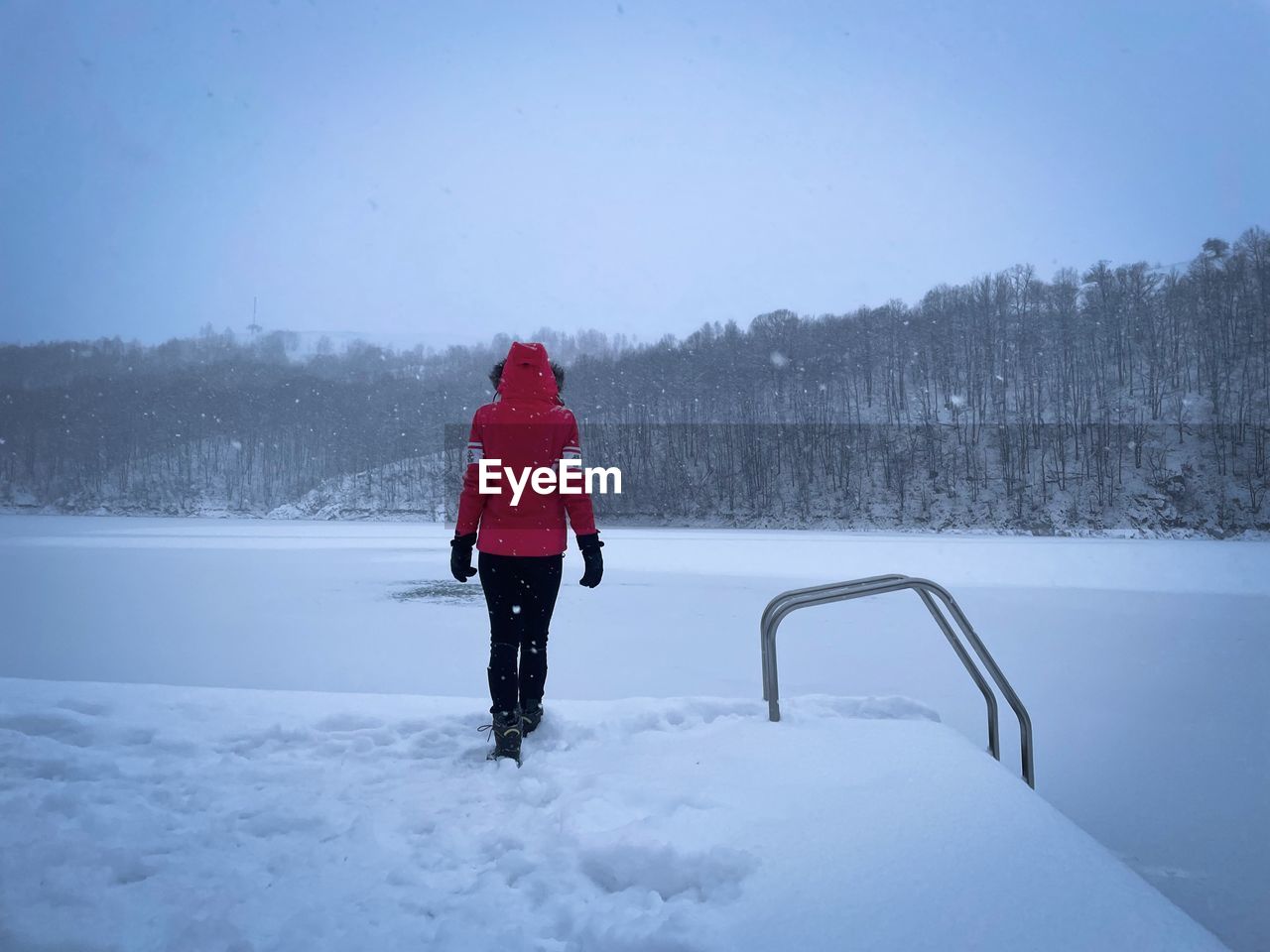 Rear view of woman in red coat standing on the pontoon near a frozen lake on a foggy winter day