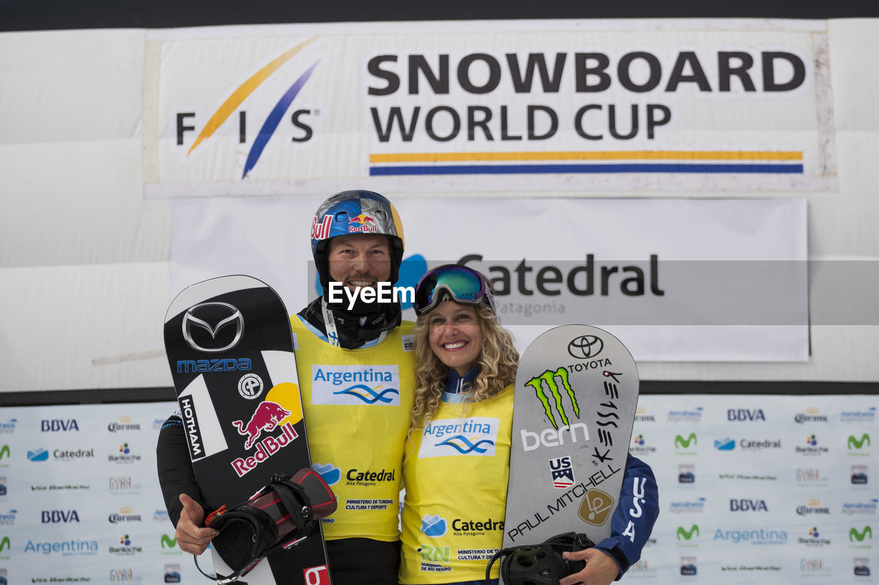 Portrait of smiling man and woman holding snowboards while standing against banner