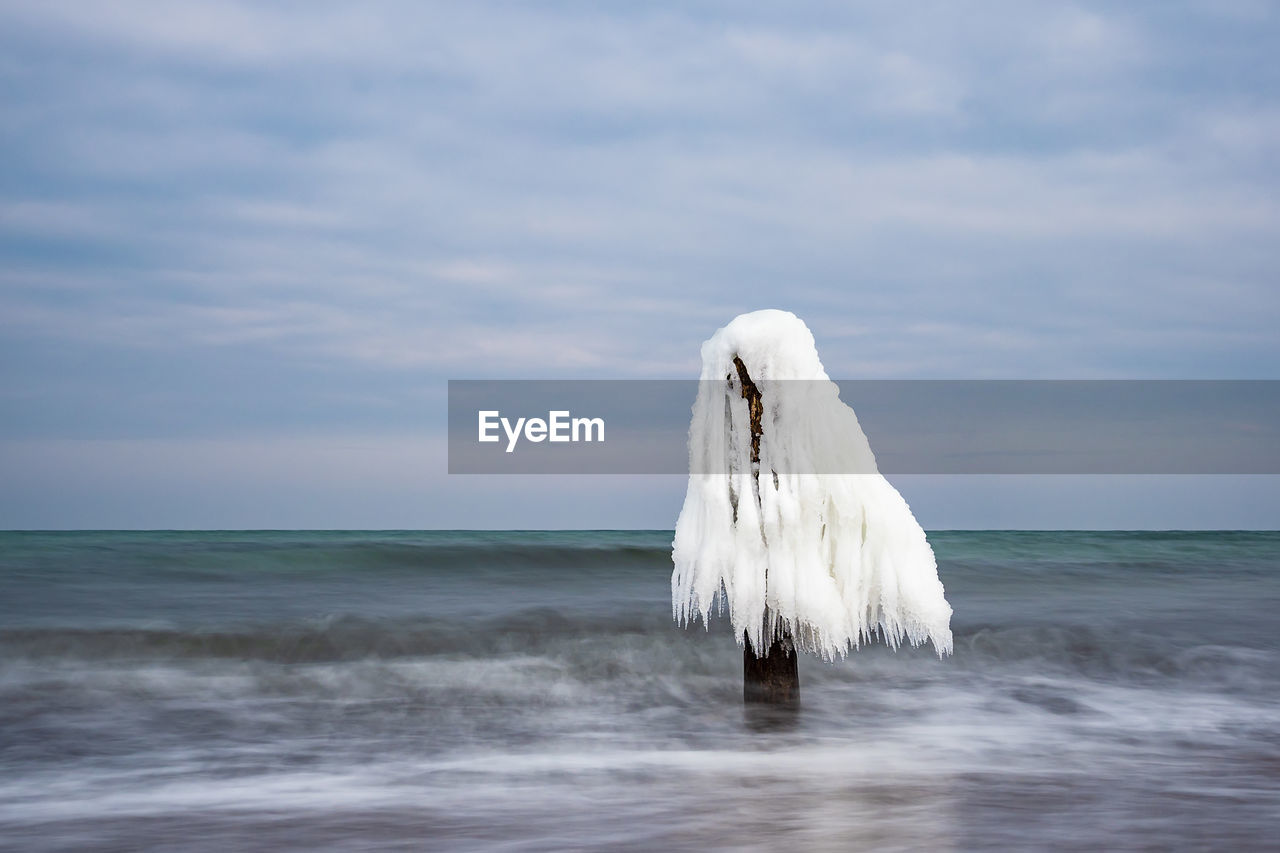 Frozen wooden post in sea against cloudy sky