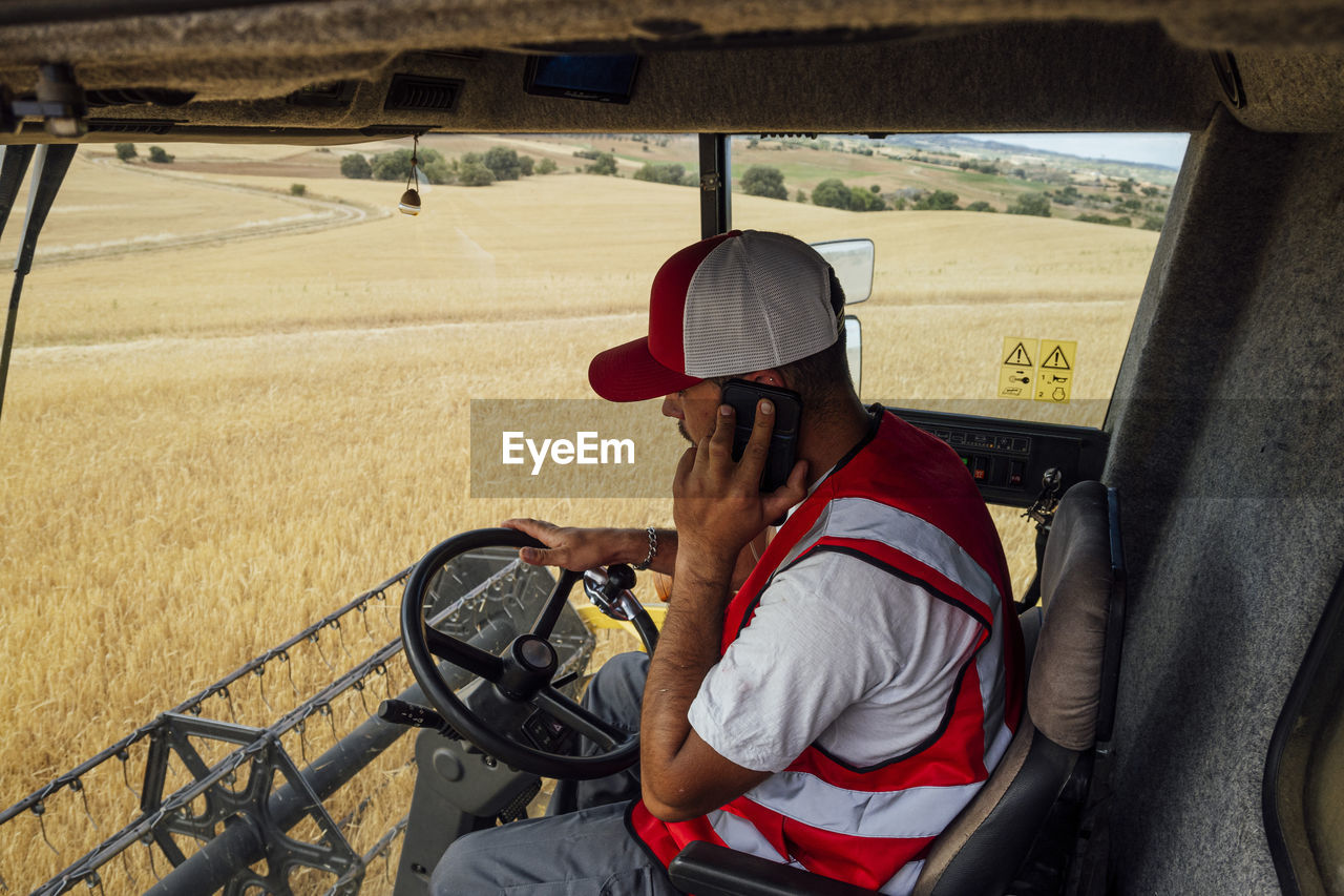 Side view of male combine operator talking on smartphone while harvesting wheat grains in professional industrial machine in agricultural field
