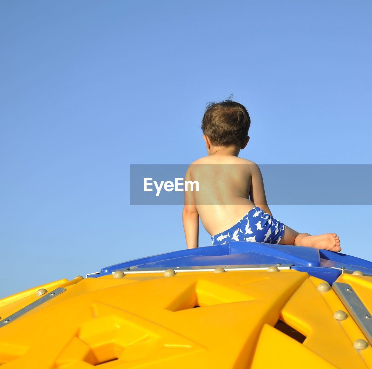 Rear view of shirtless boy sitting on play equipment against clear blue sky