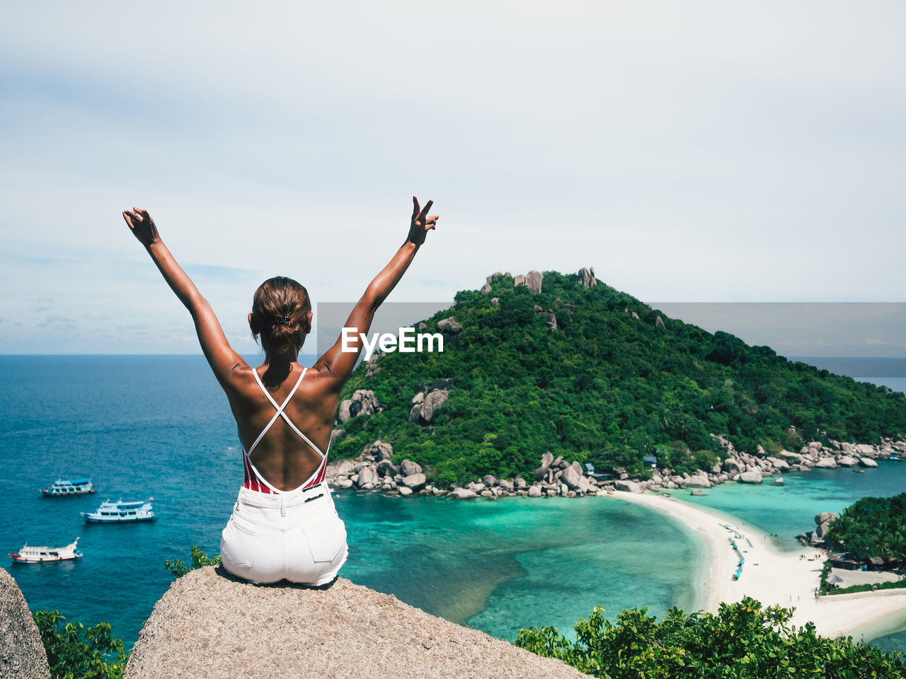 Rear view of woman with arms raised sitting on cliff against sea 