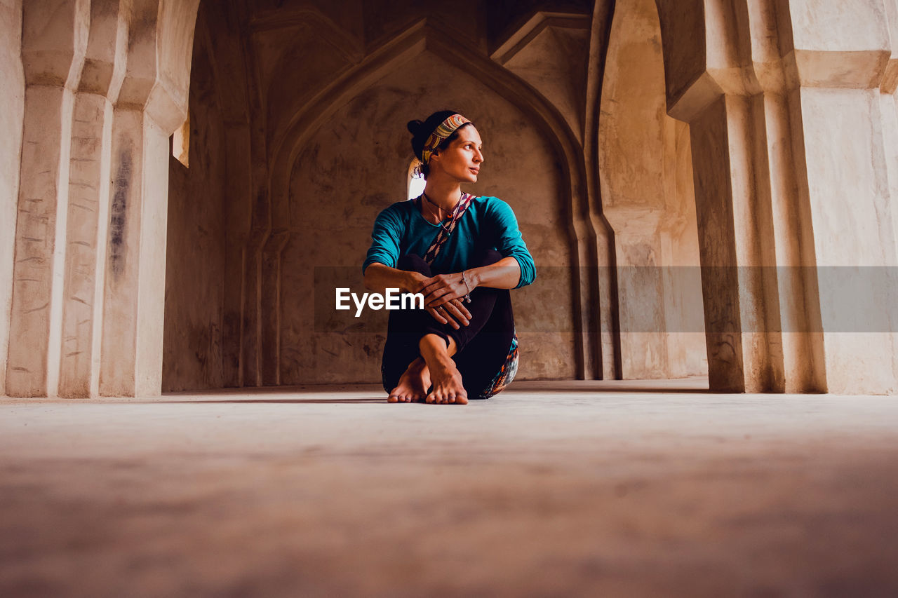 Full body of peaceful young barefooted ethnic female traveler in traditional clothes sitting on floor in ancient oriental palace in india and looking away thoughtfully