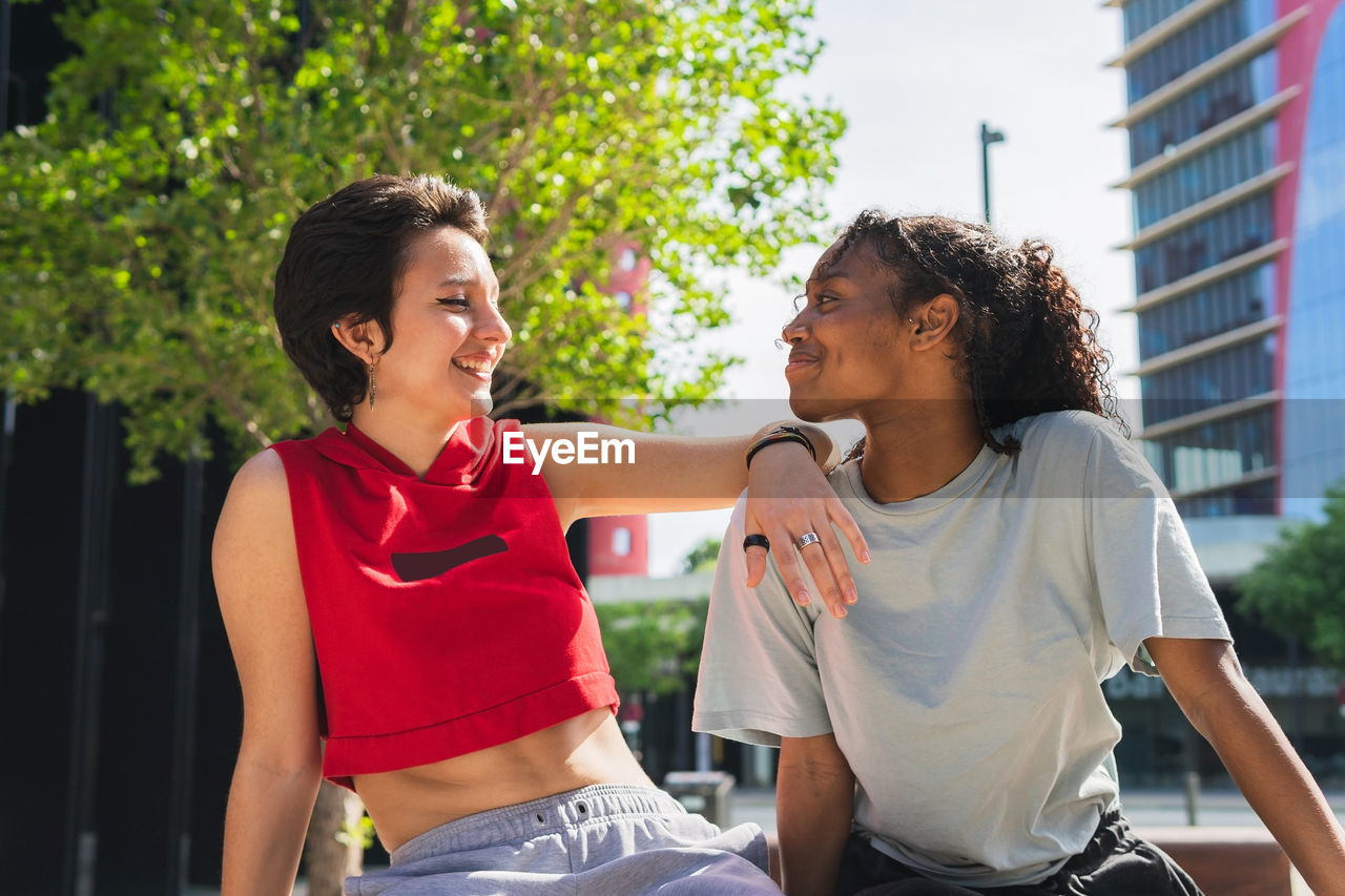 Cheerful multiracial young female friends in casual clothes looking at each other while sitting together on sunny street in city