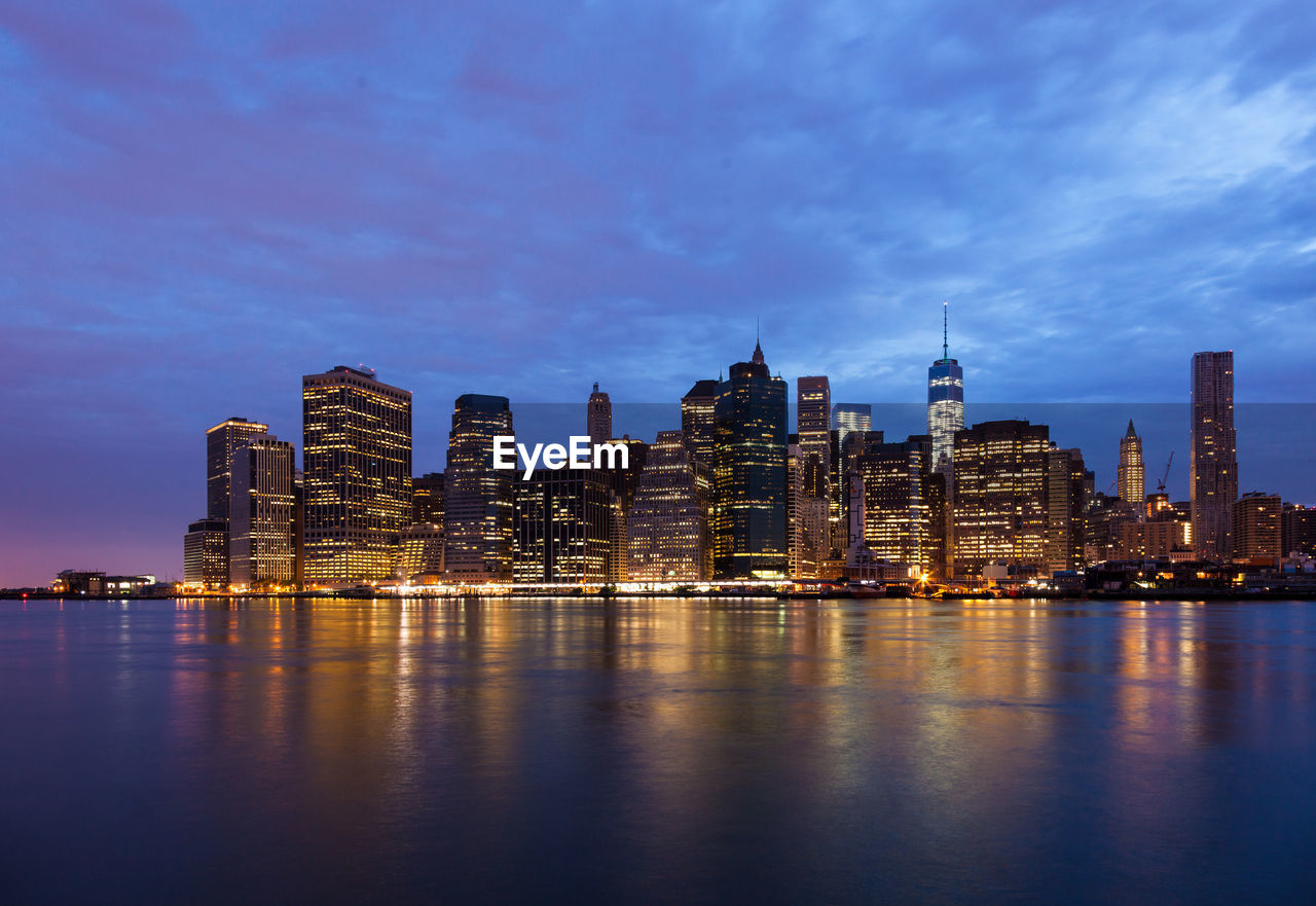 ILLUMINATED BUILDINGS BY RIVER AGAINST SKY AT DUSK