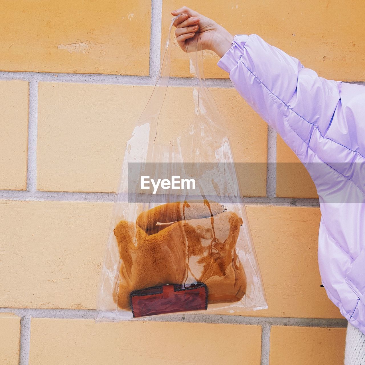 Cropped hand of woman holding warm clothing with purse in plastic bag against orange wall