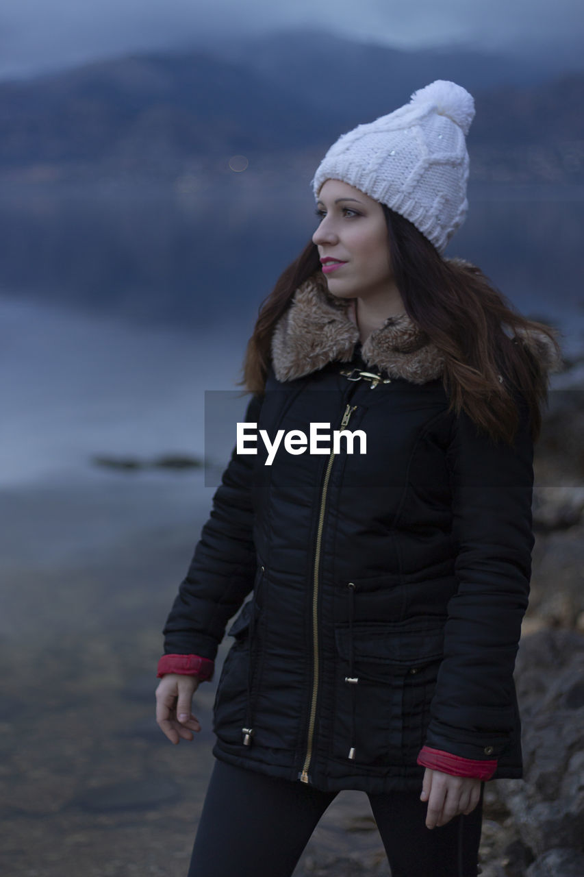 Beautiful woman looking away while standing by lake during winter