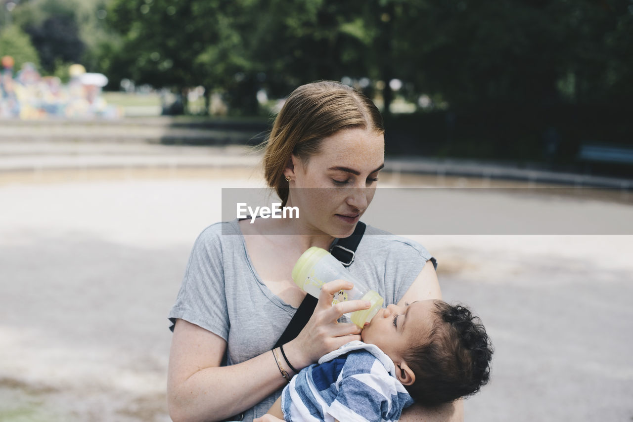 Young mother feeding baby boy from bottle at park