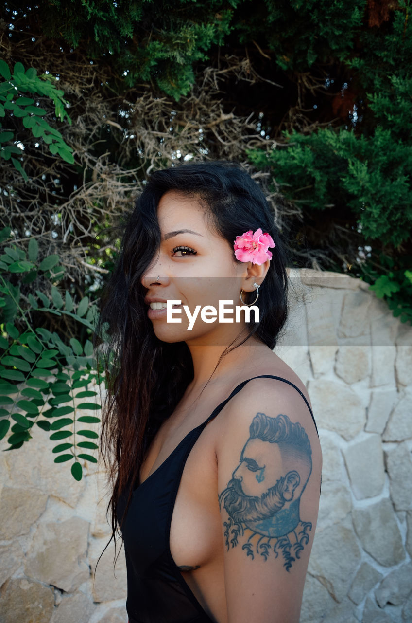 Side view portrait of woman in swimsuit with tattoo on arm standing outdoors