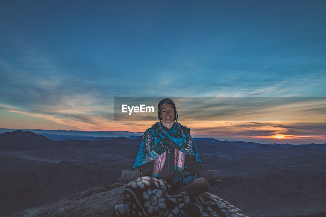 Woman meditating while sitting on rock against sky during sunset