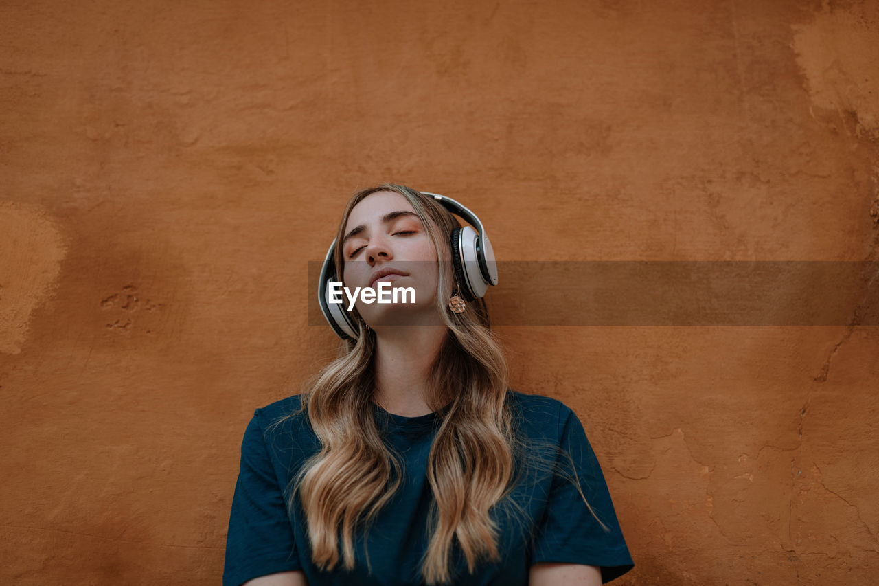 Young mindful blonde female with closed eyes listening to song in wireless headset on brown background in city