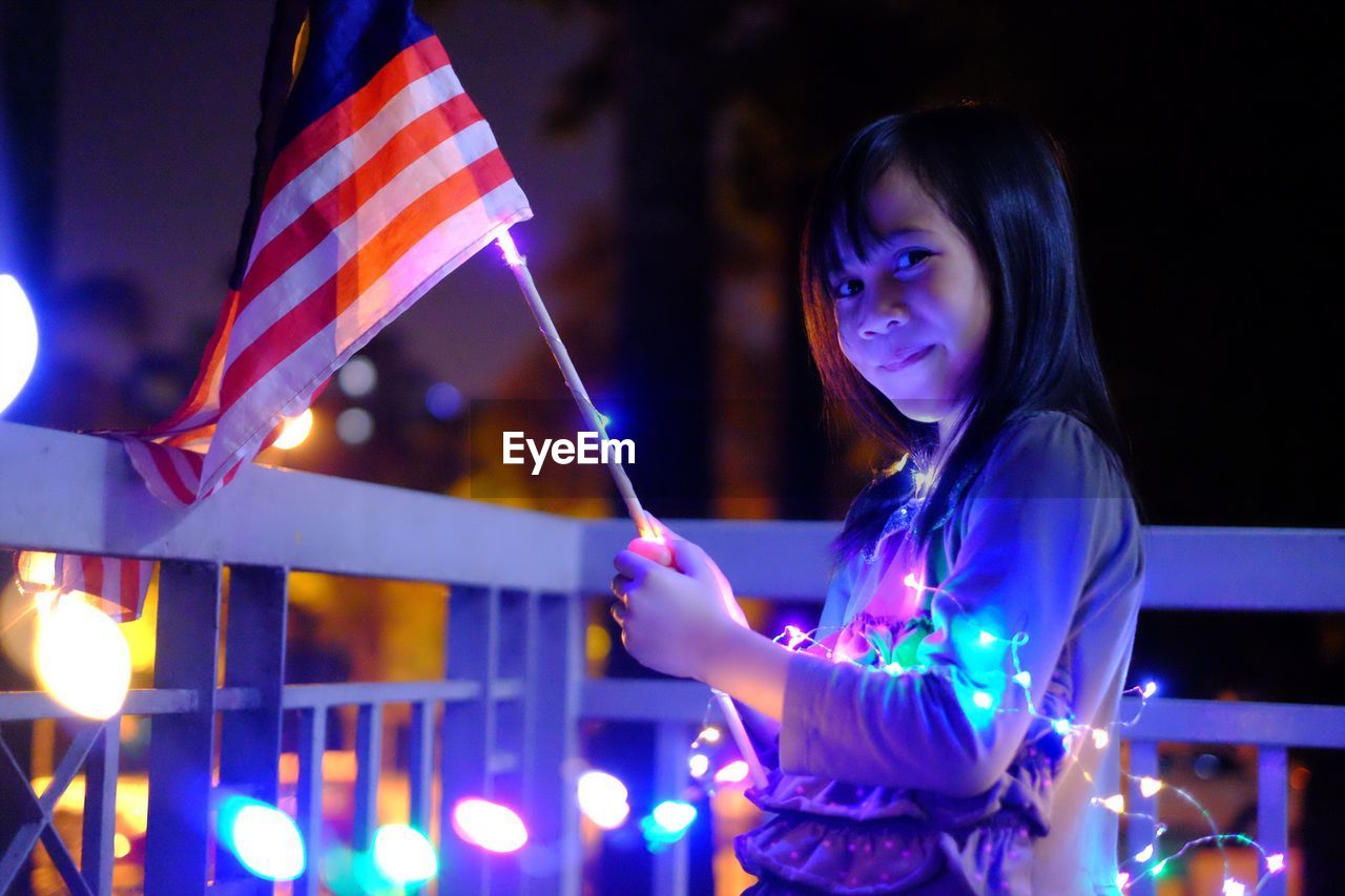 Portrait of girl with illuminated string lights and malaysian flag at night