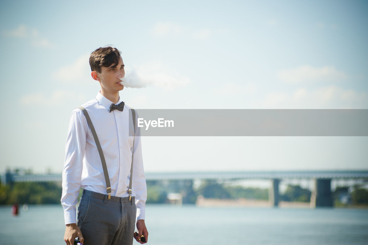 Young man smoking cigarette by river against sky