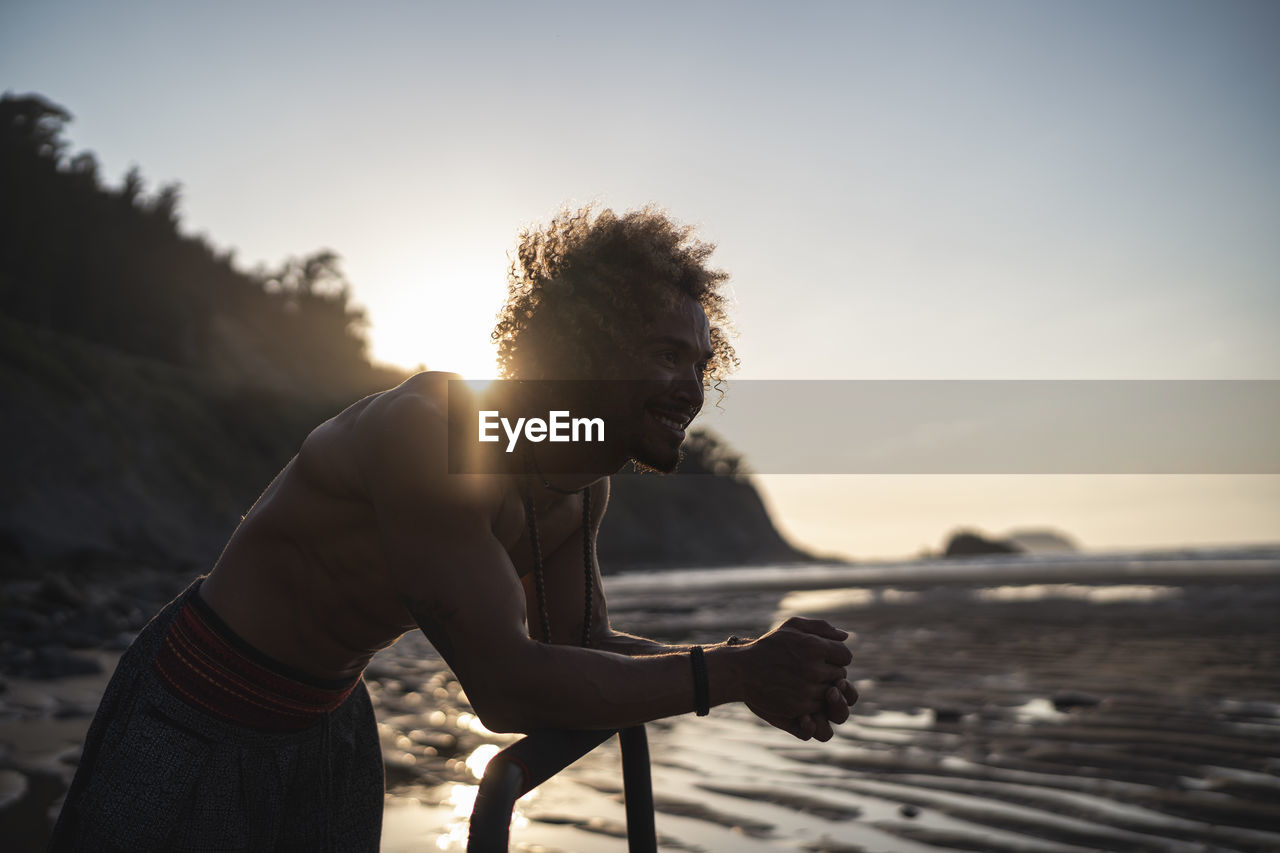 Shirtless young man looking away while leaning on parallel bars at beach