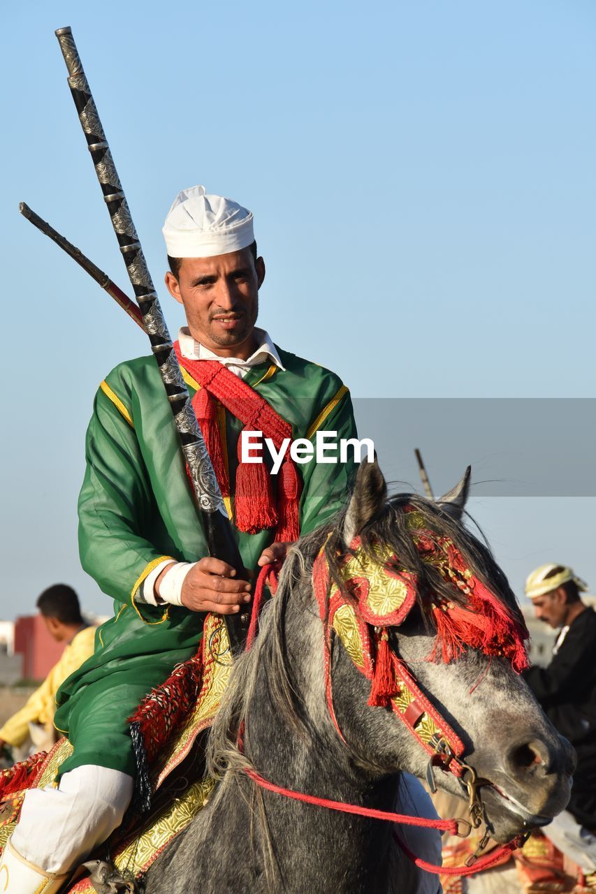 Portrait of man wearing traditional clothing riding horse against clear sky