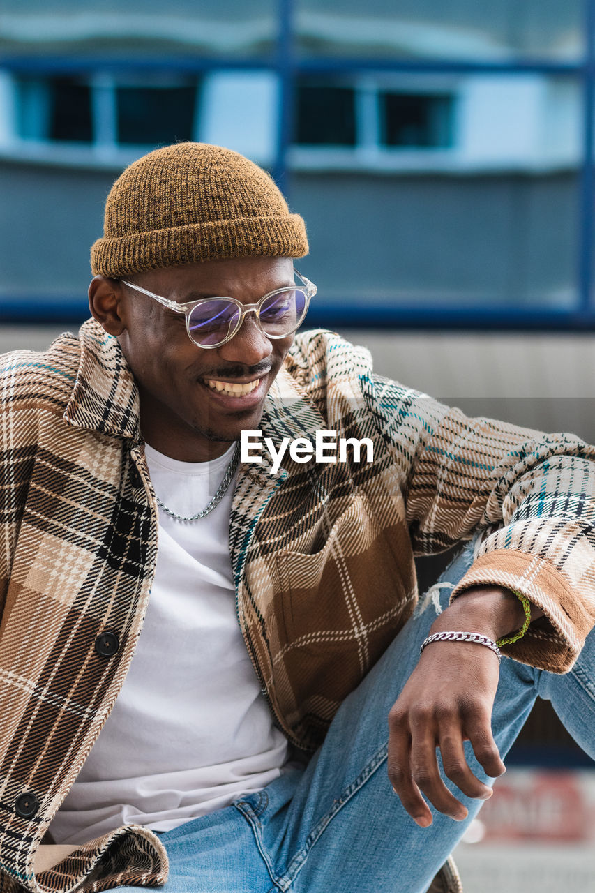 Young black guy in trendy checkered shirt frowning and looking down while chilling on street of modern city