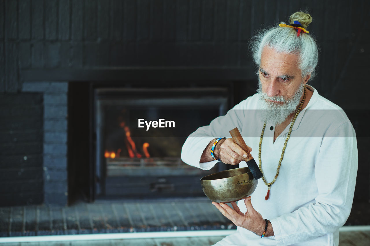 Elderly male with gray beard playing singing bowl with wooden striker while looking away during spiritual practice
