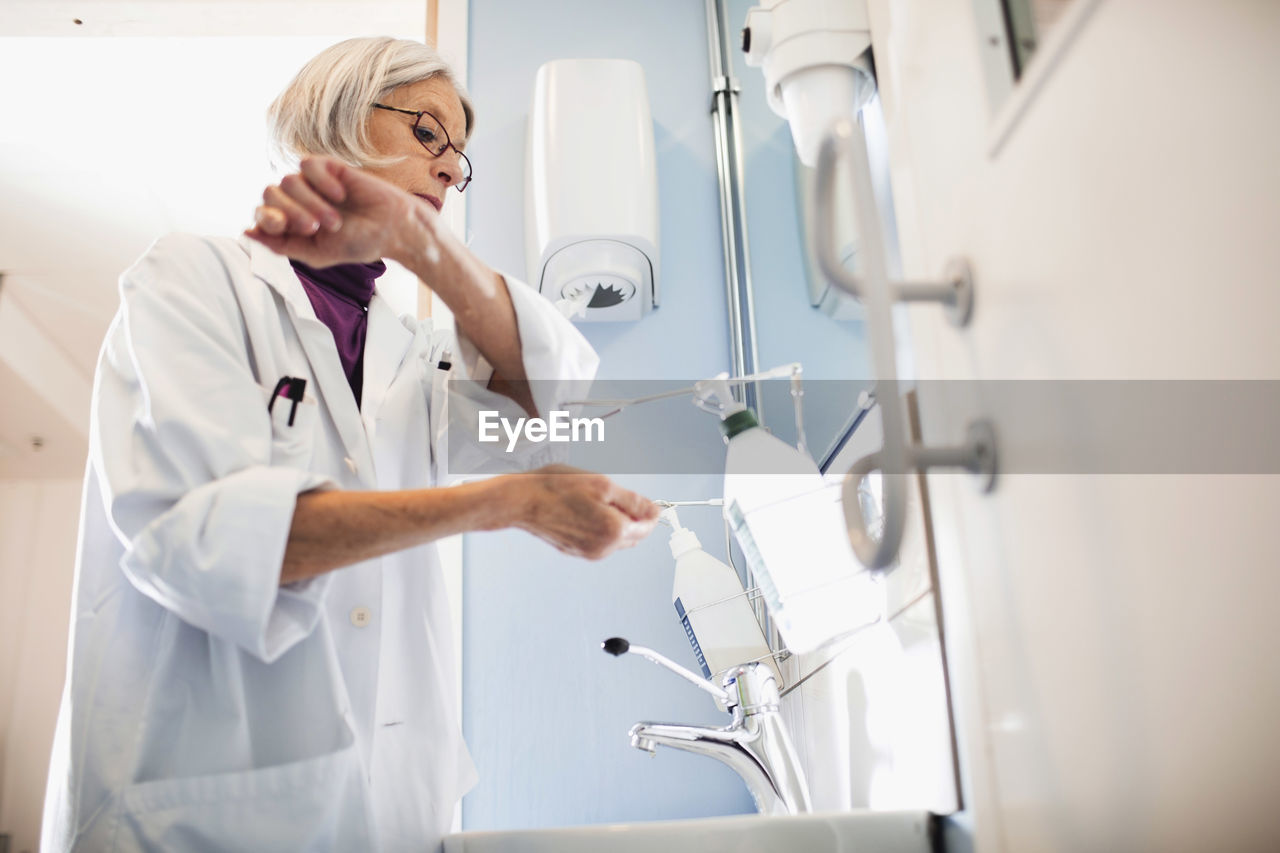Low angle view of senior female doctor using soap dispenser to wash hands in bathroom