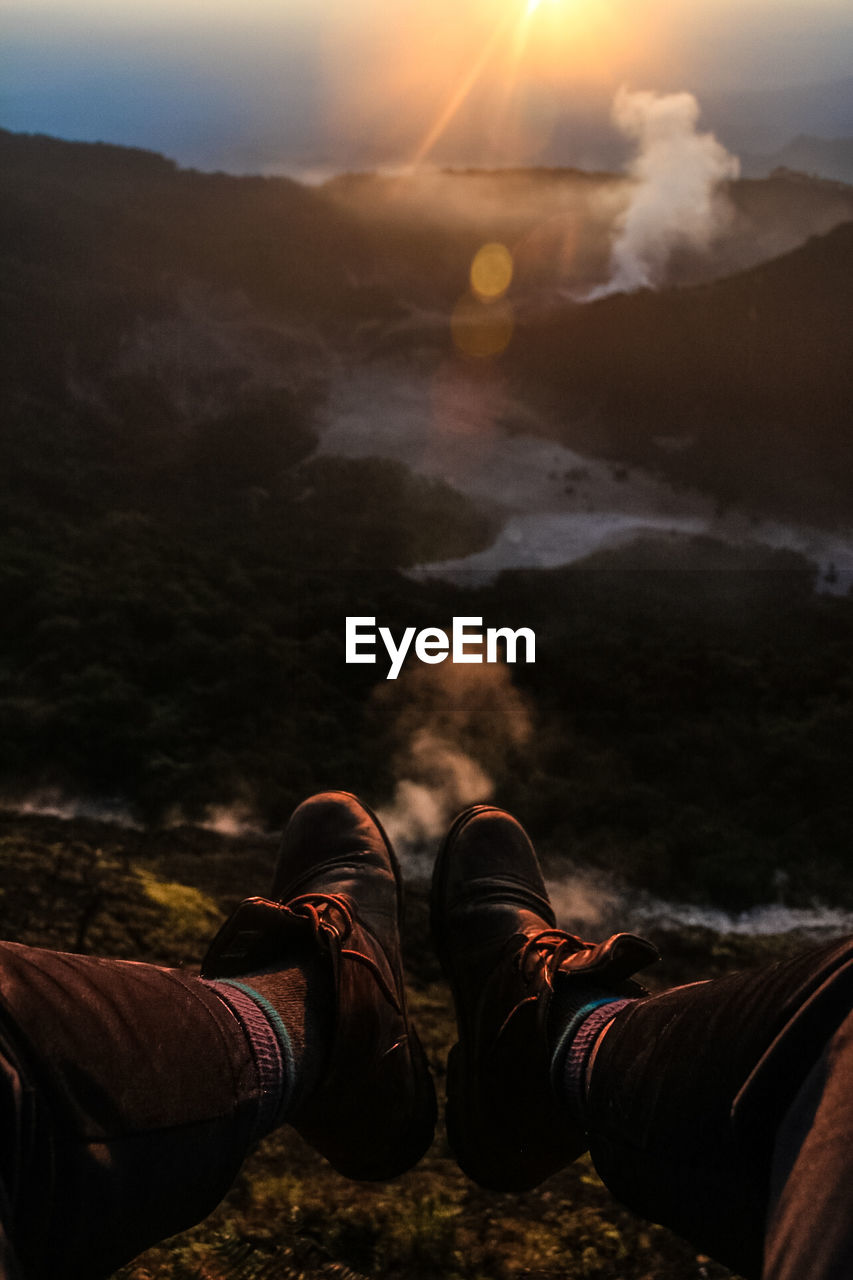 Low section of man sitting on cliff against landscape during sunset