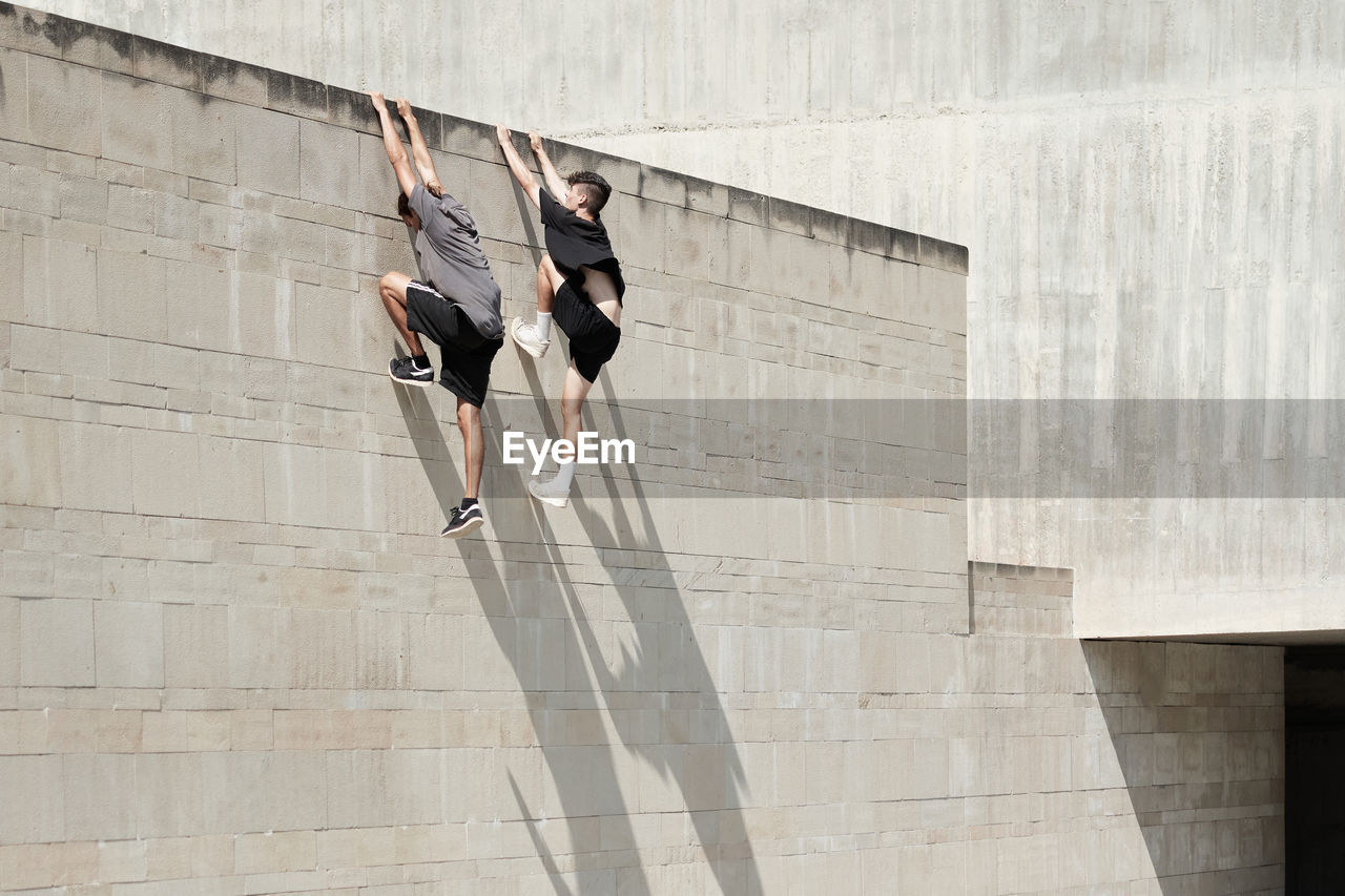 Side view of brave male friends hanging on wall of concrete building while performing risky trick and doing parkour in city