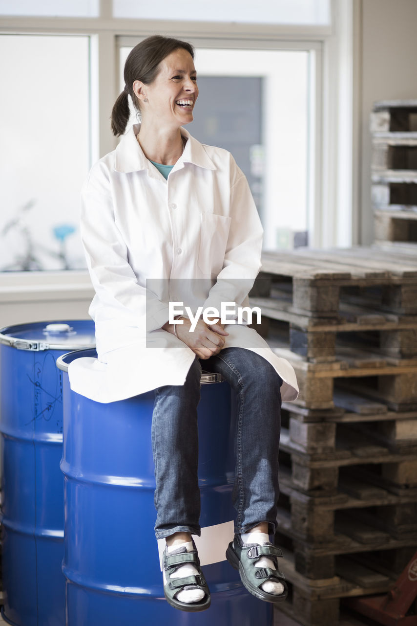 Full length of happy female engineer sitting on barrel in manufacturing plant