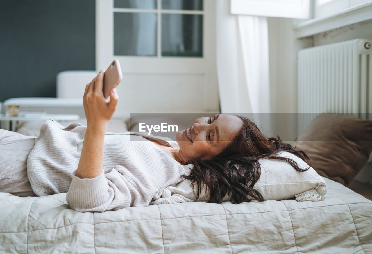 Young woman with brunette long hair in cozy knitted cardigan using mobile phone in bed at home