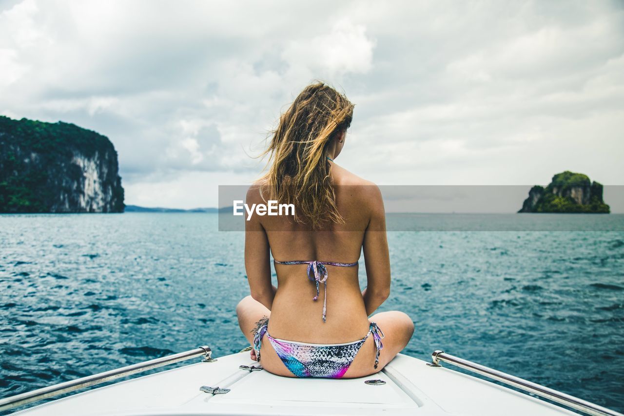 REAR VIEW OF YOUNG WOMAN IN BIKINI SITTING AGAINST SEA