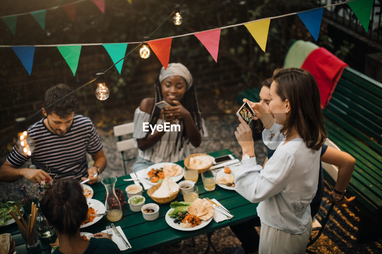 High angle view of young women photographing friends having food during dinner party in backyard