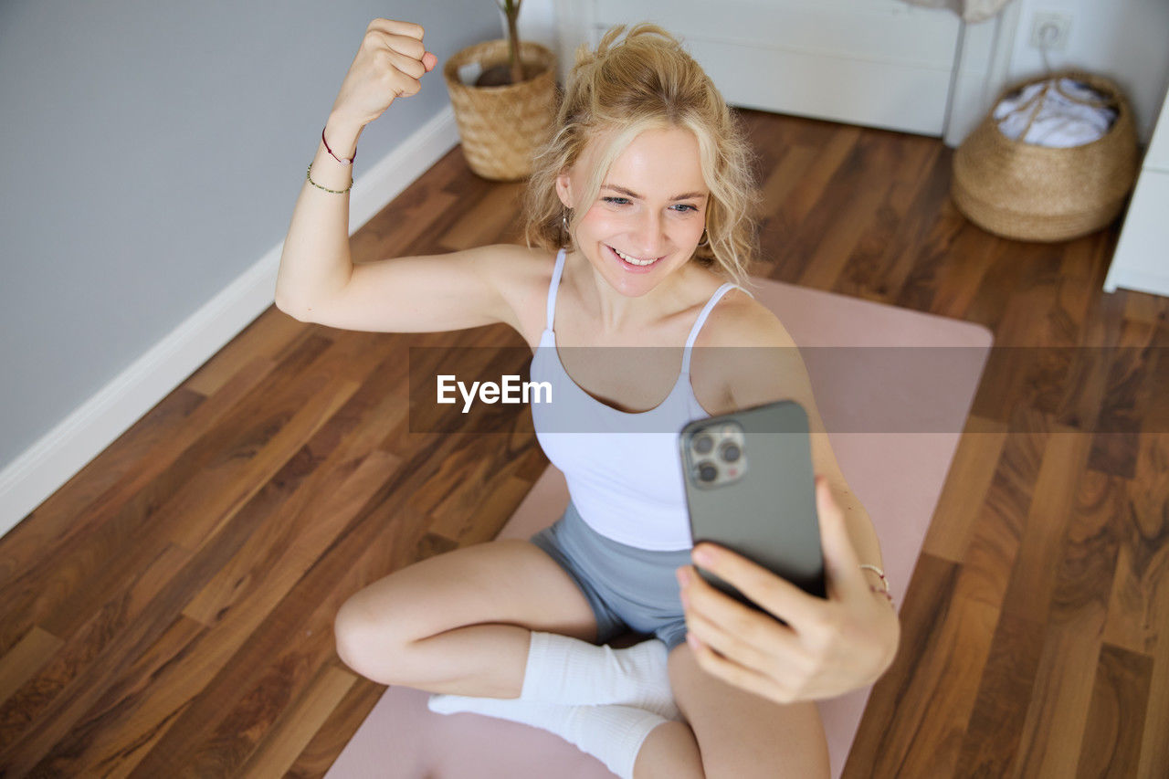 portrait of young woman using mobile phone while sitting on hardwood floor