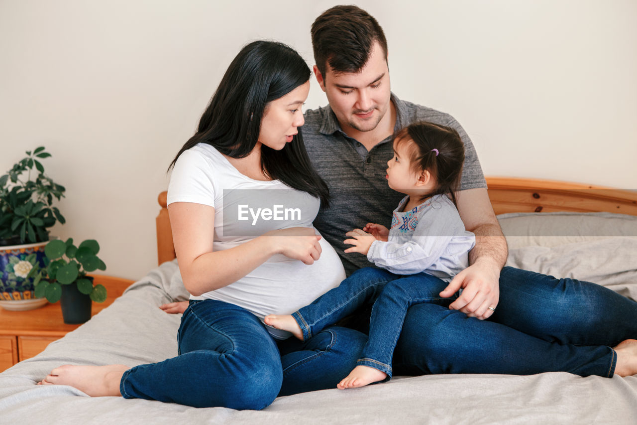 Family asian chinese pregnant woman and caucasian man with toddler girl sitting on bed at home. 