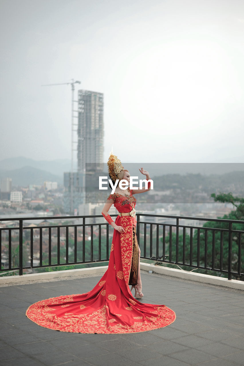 Woman with traditional custom wedding of indonesia standing by railing against sky in city