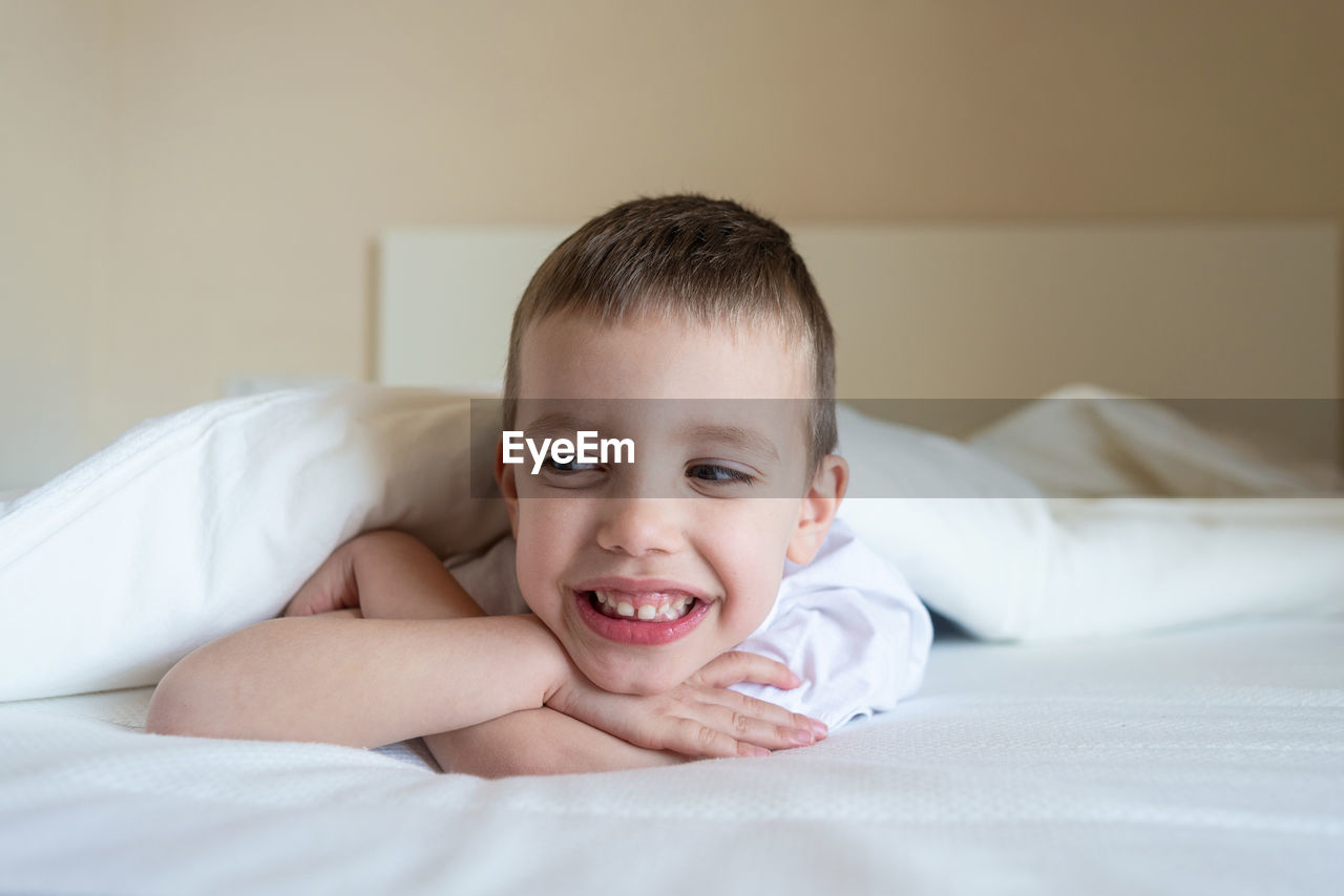 Adorable smiling kid lying down on bed under blanket, looking camera