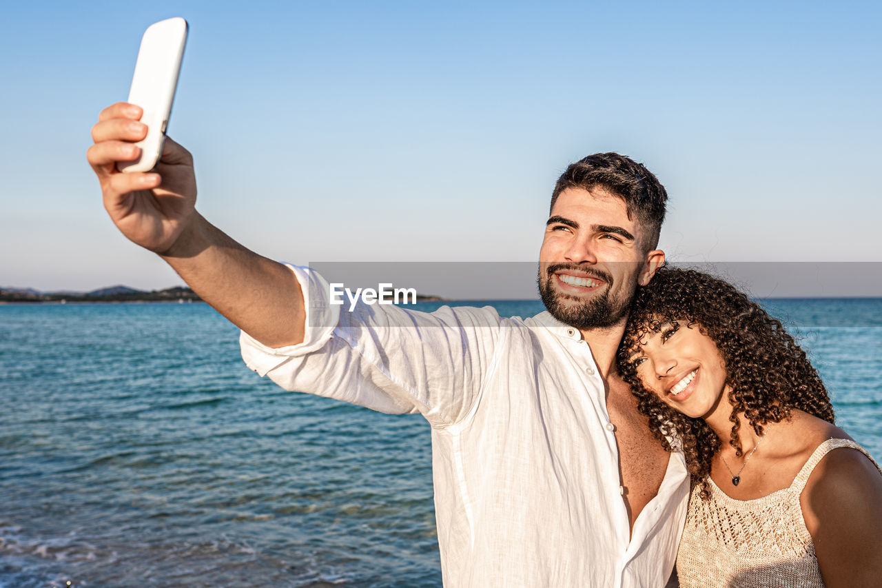 Portrait of smiling young man using mobile phone against sea