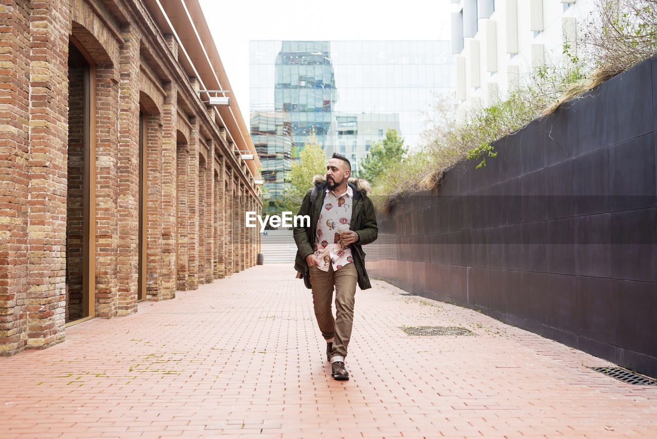 Bearded man walking across pathway holding a take away coffee